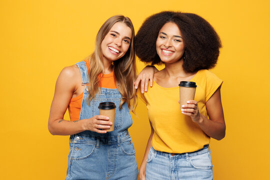 Two young happy friends buddies women wear denim casual clothes together hold takeaway delivery craft paper brown cup coffee to go isolated on plain yellow orange background studio. Lifestyle concept.