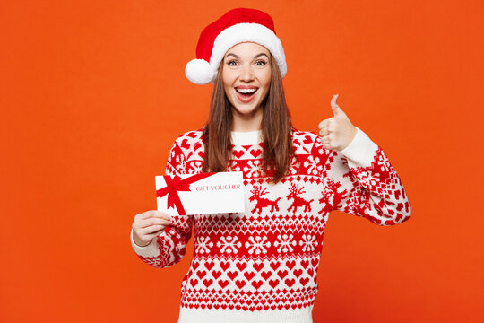 Merry young woman wear red warm Christmas sweater Santa hat posing hold gift coupon voucher card for store show thumb up isolated on plain orange background Happy New Year celebration holiday concept