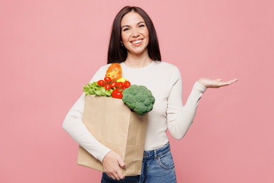 Young happy woman wear white casual clothes hold brown craft bag for takeaway mock up with food products point hand aside show empty palm isolated on plain pink background. Delivery service from shop.