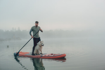 young man with tattooed arms on a sup board with his samoyed dog at the lake during sunset