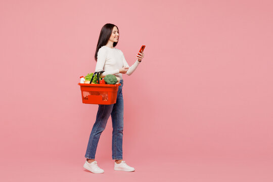 Full body side view young happy woman wearing white casual clothes hold orange basket bag with food products use mobile cell phone walk go isolated on plain pink background Delivery service from shop