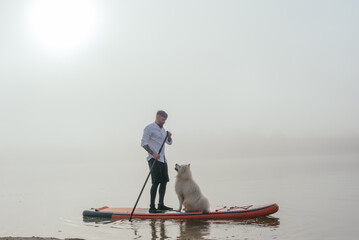 young man with tattooed arms on a sup board with his samoyed dog at the lake during sunset