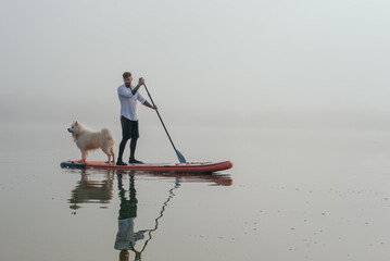 young man with tattooed arms on a sup board with his samoyed dog at the lake during sunset