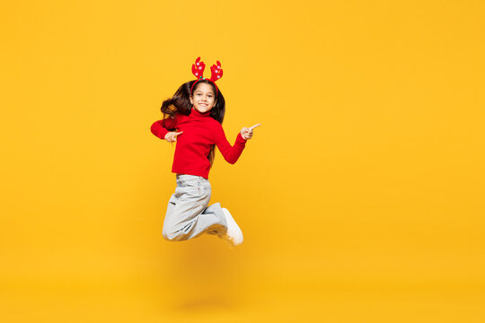 Full body merry little girl wear red clothes fun decorative deer horns on head posing jump high point finger aside on area isolated on plain yellow background Happy New Year Christmas holiday concept