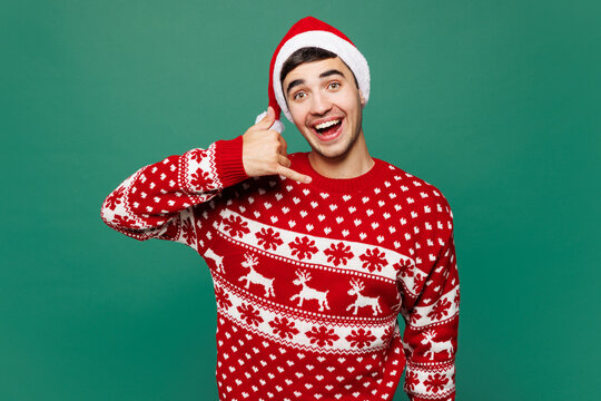 Merry young man wear red warm cozy Christmas sweater Santa Claus hat posing doing phone gesture like says call me back isolated on plain green background. Happy New Year celebration holiday concept.
