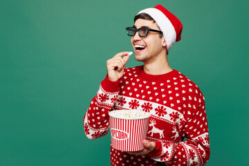 Merry young man wears red cozy Christmas sweater Santa Claus hat 3d glasses watch movie film hold bucket of popcorn in cinema posing isolated on plain green background. Happy New Year holiday concept.