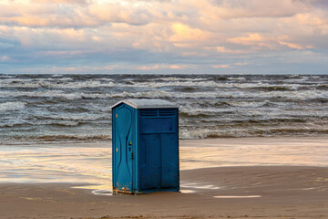 Blue portable restroom stands on sandy beach near the ocean during sunset hours