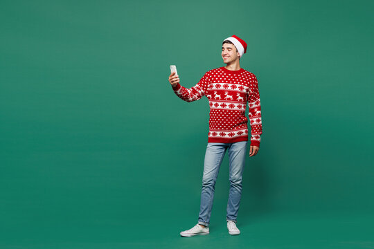 Full body merry young man wear red warm cozy Christmas sweater Santa Claus hat posing hold in hand use mobile cell phone chat online isolated on plain green background. Happy New Year holiday concept.
