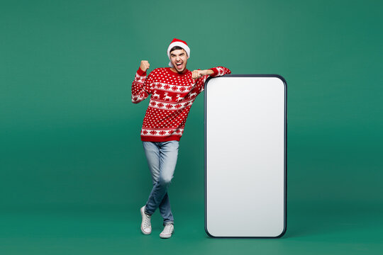 Full body merry young winner man wear red Christmas sweater Santa Claus hat posing big huge blank screen mobile cell phone area smartphone isolated on plain green background. Happy New Year concept.