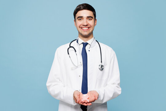 Young male doctor man wear white medical gown suit work in hospital clinic office hold in hand pink ribbon isolated on plain blue background studio Healthcare medicine breast Cancer Awareness concept.