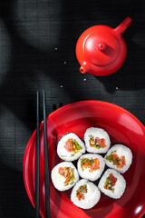 A red plate with maki rolls, chopsticks, red teapot on a black bamboo mat in soft sunlight.