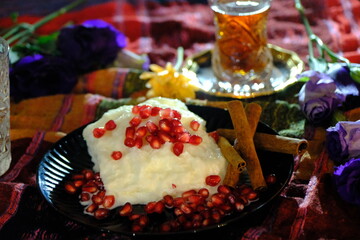 Ramadan dessert: Güllaç. Topped with pomegranate and cinnamon sticks. Tea in a crystal glass. Silver tea saucer. Purple roses and yellow daisies. Iftar table. Turkish milk desserts.
