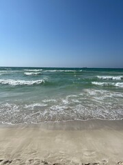 A calm sandy beach with gentle turquoise waves rolling to shore under a bright blue sky — a serene coastal view perfect for relaxation, travel, and nature backgrounds.