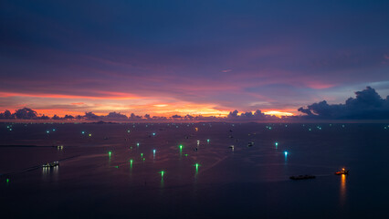 twilight time over the sea, The sunset sky is filled with deep purple, orange, and pink hues. Bright green lights from fishing and other boats twinkle on the water,