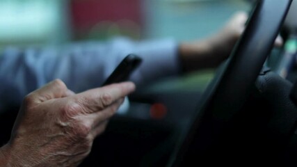 Handling your phone while driving. Close-up of old man hand holding a phone and steering wheel of a car while driving. Closeup inside vehicle of hand holding smart phone and steering wheel.