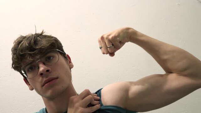 A young caucasian man with glasses lifts his t-shirt sleeve and flexes his bicep muscle, showing strength and pride.