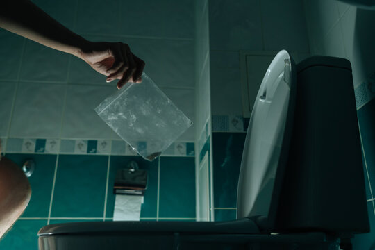 Close-up cropped shot of male hand holding plastic bag containing illicit pills hovering above toilet bowl, symbolizing drug addiction recovery and substance disposal process in dark bathroom.
