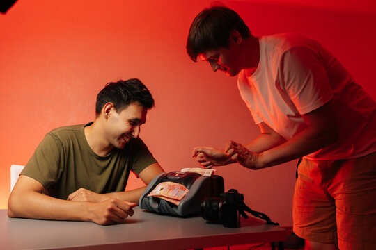 Two cheerful male content creators sitting at workspace, counting earnings near professional cameras under red light, showcasing successful digital entrepreneurship, studio shot, isolated background