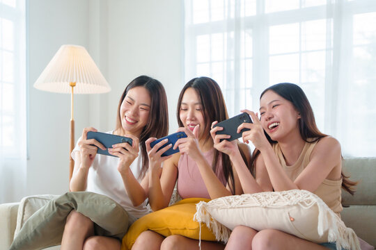 Three Asian women friends playing mobile games with smartphone and laughing together at home, showing friendship and fun lifestyle.