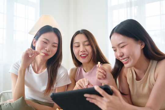 Three Asian women friends sitting together watching movie and shopping online on tablet, enjoying relaxing weekend at home.