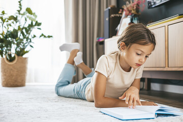 Young girl lying on a floor in a modern living room reading a book. Concept of independent childhood learning and leisure