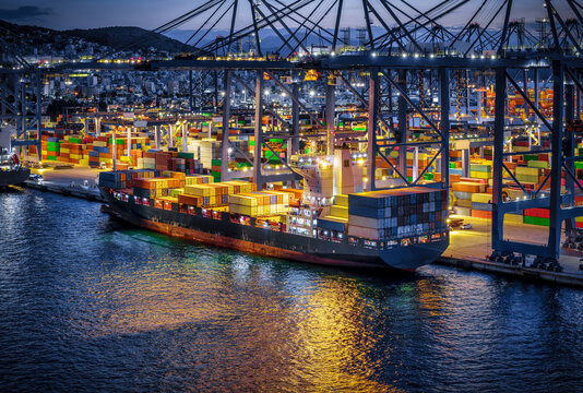 Aerial view of a container cargo ship being unloaded and loaded by cranes in a commercial harbour dock during evening time
