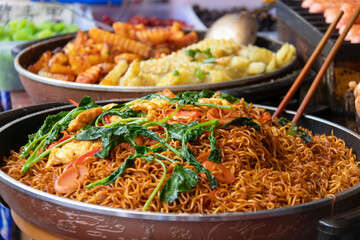 Stir fried noodle dish with greens, egg and sliced sausage served in market pan, savory street food scene with warm colors