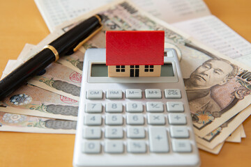 計算機の上に置かれた家の木製おもちゃ、銀行通帳、ペンと一万円札の紙幣 お金のイメージ
