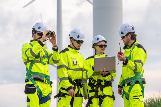 Four people in yellow safety gear are standing around a laptop. One of them is holding a remote control. Engineer team in safety harnesses and PPE, preparing for wind turbine maintenance.