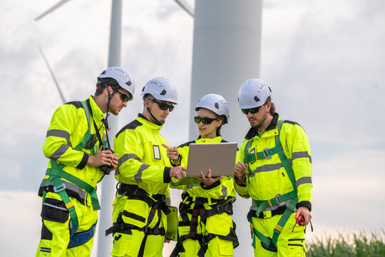 Four people in yellow safety gear are looking at a laptop. One of them is wearing a harness. Engineer team in safety harnesses and PPE, preparing for wind turbine maintenance.