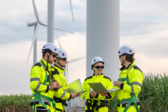 Four people wearing yellow and green safety gear are standing around a wind turbine. Engineer team in safety harnesses and PPE, preparing for wind turbine maintenance.