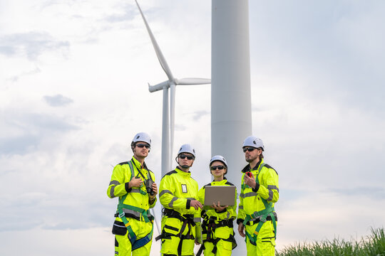 Four men in yellow safety gear are standing in front of a wind turbine. One of them is holding a laptop. Engineer team in safety harnesses and PPE, preparing for wind turbine maintenance. - Powered by Adobe