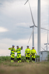 Four men in yellow and green safety gear are standing in a field. They are looking at a map. Group of engineers with PPE, collaborating and walking at wind turbine field, concept of achievement.