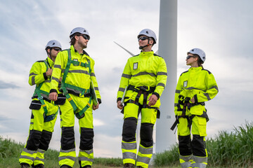 Four people in yellow and black safety gear. One of them is wearing a harness. Group of engineers (four people) with PPE, collaborating and walking at wind turbine field, concept of achievement.