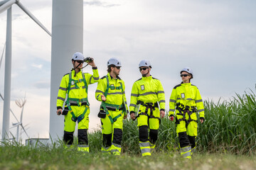 A group of four workers in bright yellow and green safety gear are walking through a field. Group of engineers with PPE, collaborating and walking at wind turbine field, concept of achievement.
