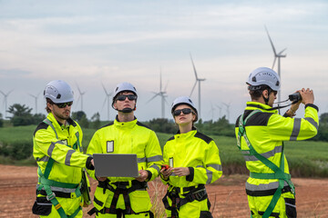 Four people in yellow safety gear are standing in a field. One of them is holding a laptop. Technicians in PPE discussing blueprint and inspecting wind turbines, planning for maintenance.