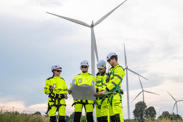 A group of people are standing in front of a wind turbine. They are wearing yellow. Technicians in PPE discussing blueprint and inspecting wind turbines, planning for maintenance.