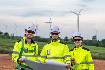Three people in yellow jackets are standing in a field. One of them is holding a map. Technicians in PPE discussing blueprint and inspecting wind turbines, planning for maintenance.
