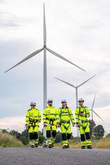 A group of four workers in bright yellow and black safety gear are walking near a large wind turbine. Technicians in PPE discussing blueprint and inspecting wind turbines, planning for maintenance.