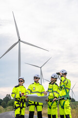 Four people wearing yellow and black safety gear are standing in front of a wind turbine. Technicians in PPE discussing blueprint and inspecting wind turbines, planning for maintenance.