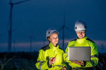 Two workers in safety gear are looking at a laptop computer. One of them is holding a remote control. Technician team in safety harnesses, inspecting wind turbines with technology at twilight.