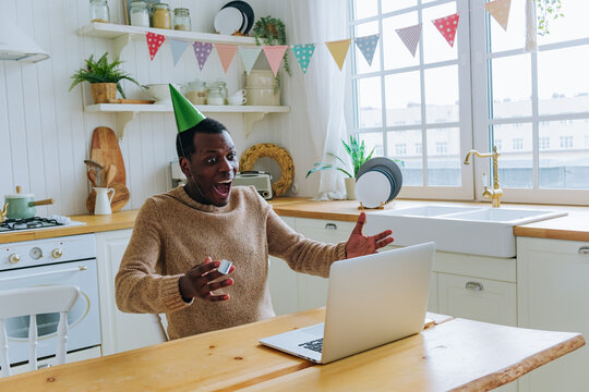 African american man in a party hat celebrating his birthday online via a laptop video call, joyfully reacting to a virtual surprise or message from friends and family in his bright kitchen