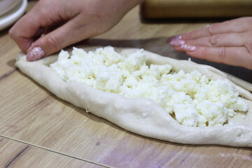 Woman folding dough into a boat shape, shredded suluguni cheese on the dough, preparing a Georgian cuisine dish, khachapuri. Close-up.