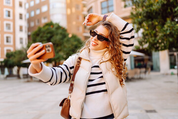 Cheerful woman in sunglasses blogs on phone while strolling along sunny city streets. Beautiful...
