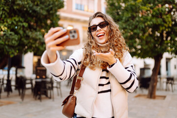 Cheerful woman in sunglasses blogs on phone while strolling along sunny city streets. Beautiful...