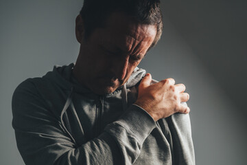 Mature man in low key light holding shoulder in severe pain, concept of injury, muscle strain, or chronic joint problem.