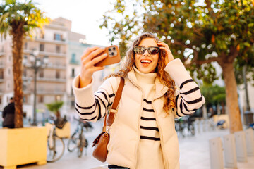 Cheerful woman in sunglasses blogs on phone while strolling along sunny city streets. Beautiful...