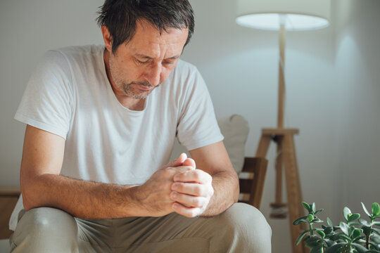 Lonely casual male with a sorrowful, regretful expression sitting indoors, feeling down and lost in thought in a dim cozy living room
