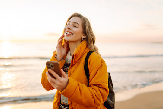 Portrait of cheerful woman with curly hair strolling along the beach at sunset, holding mobile phone. Young traveler with phone enjoys seascape. Concept of hiking and adventure.