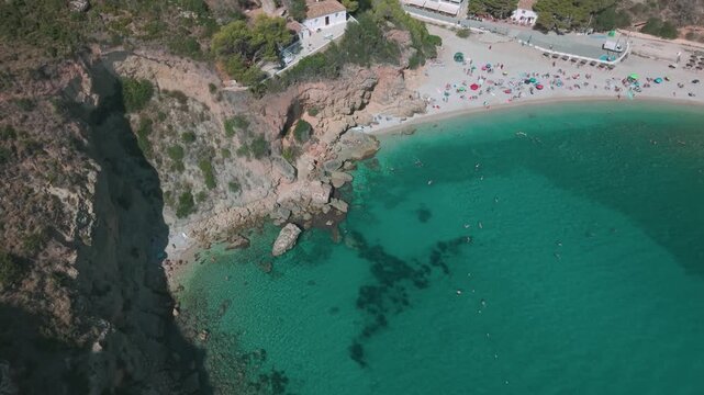 Top-down aerial view of Cala Granadella beach with people swimming in clear turquoise water and sunbathing on the shore, Javea, Spain.